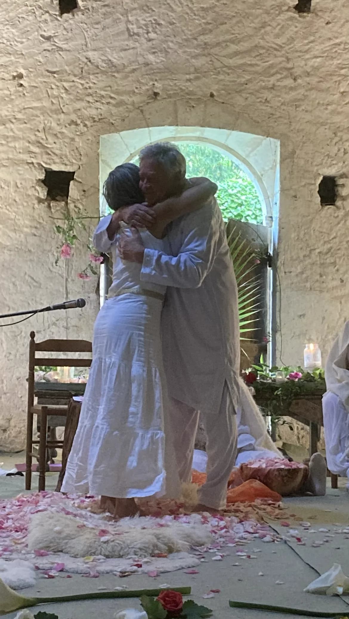 Deux personnes s'étreignent lors d'une cérémonie, dans une chapelle de pierre, sur un tapis de pétales