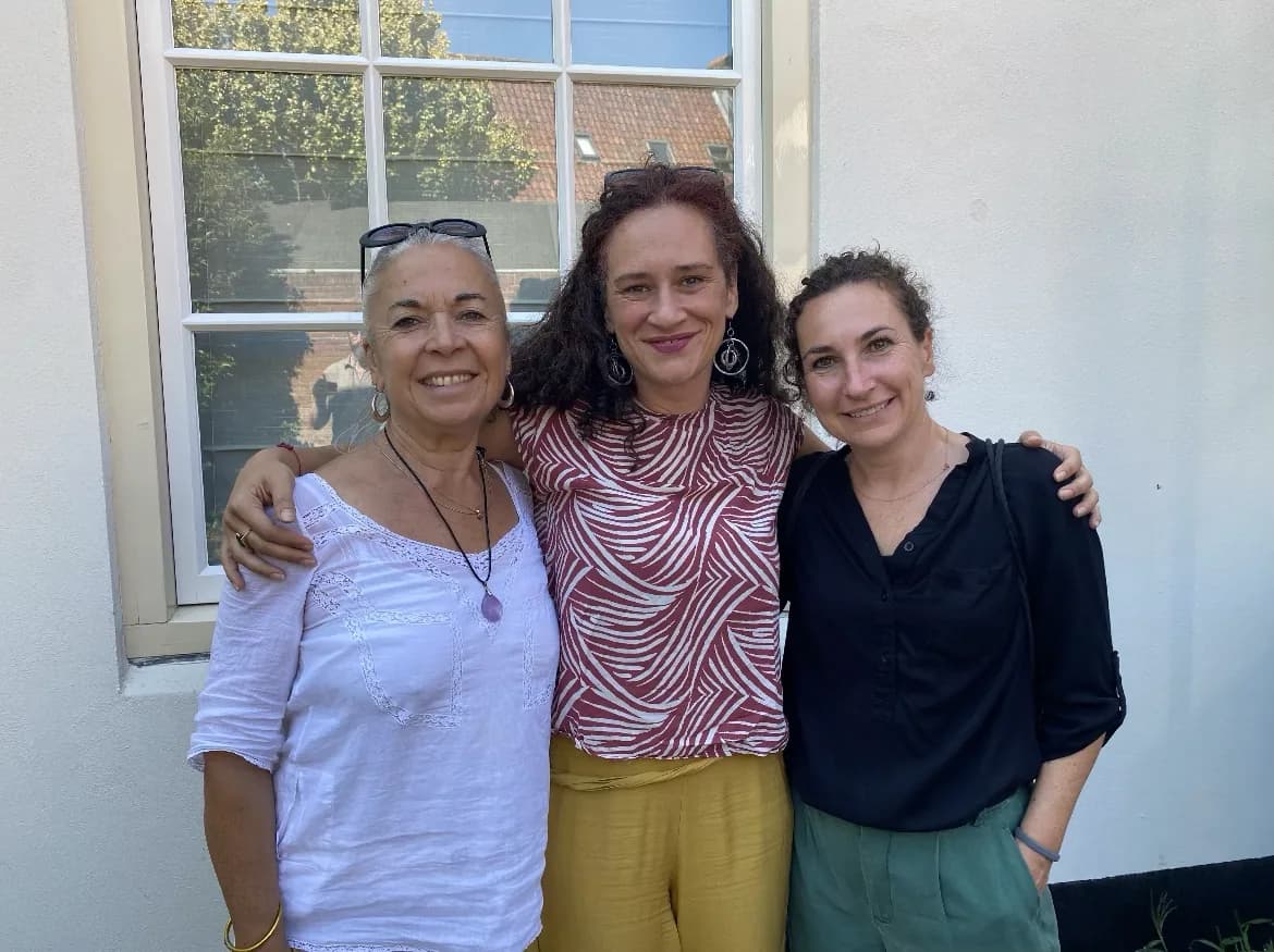 Trois femmes souriantes, bras sur les épaules, devant une fenêtre